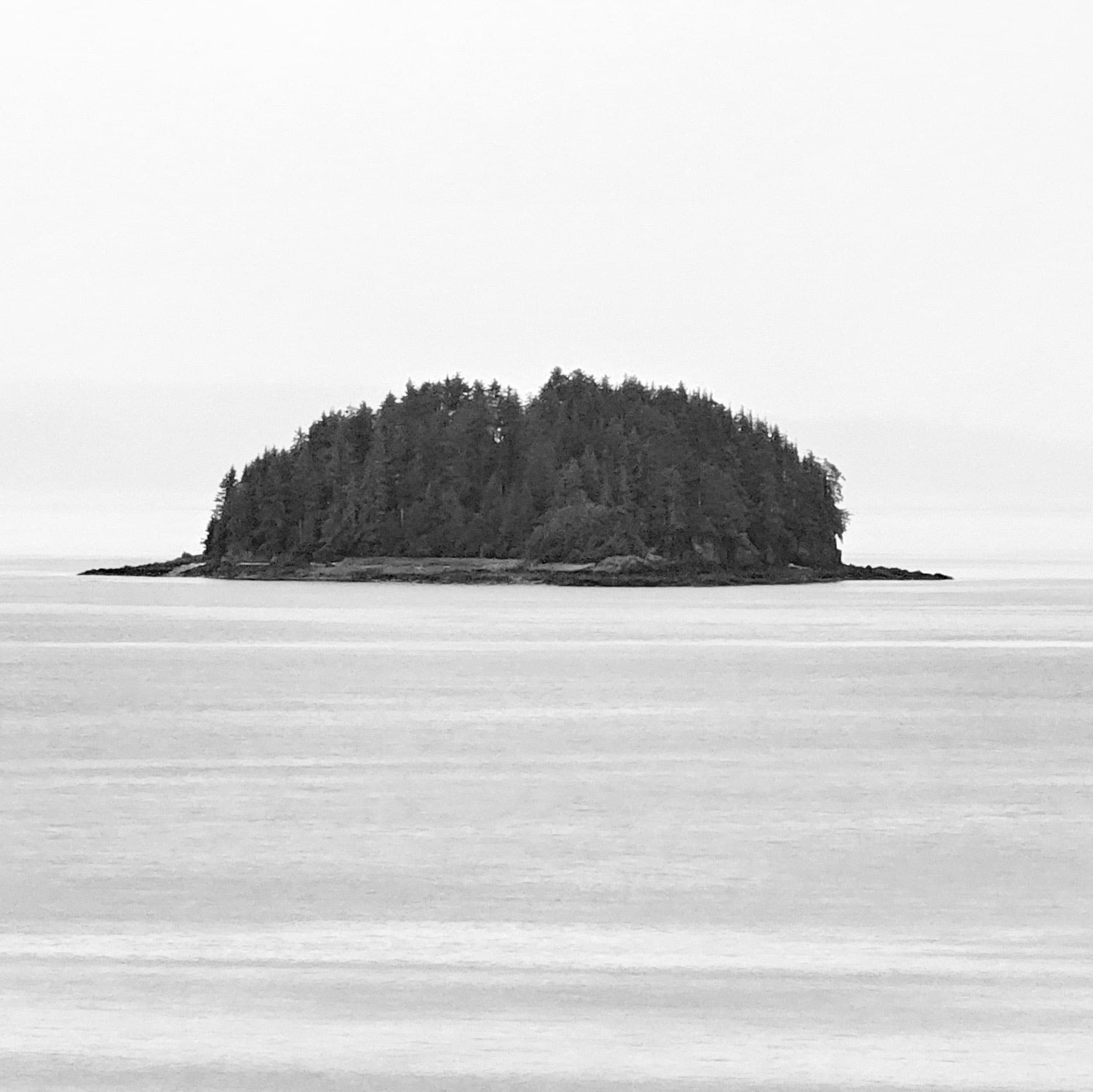 A small dark forested island floating where pale sea and sky merge into one field of mist, Inside Passage, Alaska.